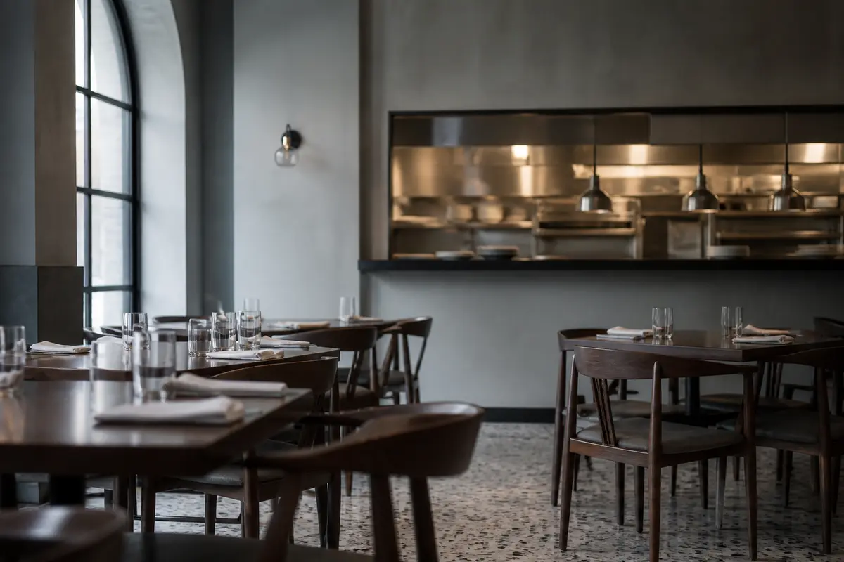 Restaurant dining room set before service: laid tables, dark wood chairs and the lit kitchen pass at the back.